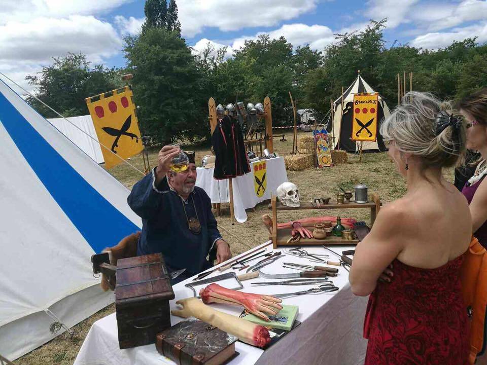 Un membre de la troupe présente des outils d'époque devant les tentes et les boucliers aux couleurs des Ganecoux