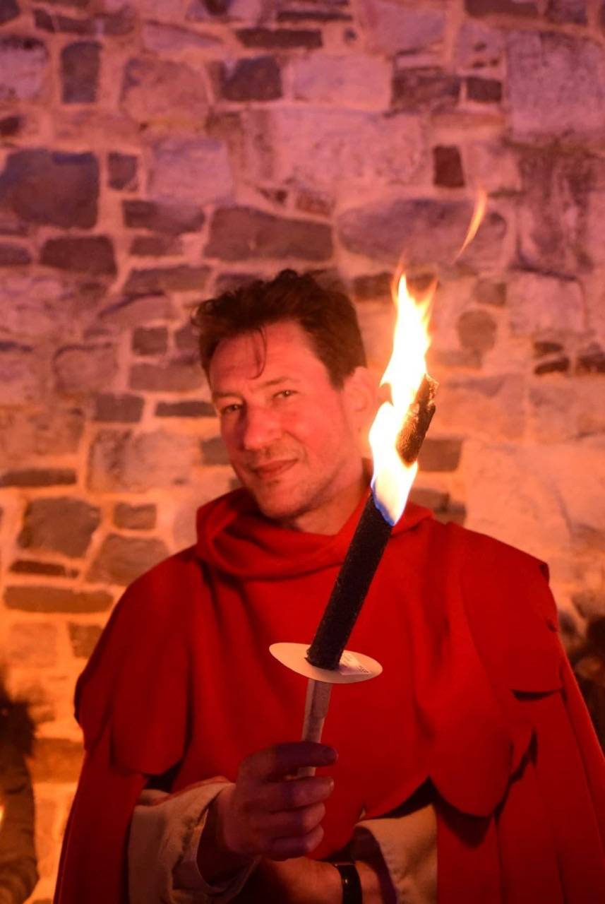Un homme en tenue médiévale rouge tient un flambeau dans la nuit